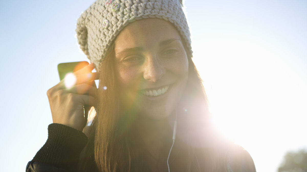 Junge Frau mit Mütze hört Musik, lächelt und genießt den sonnigen Tag im Freien.