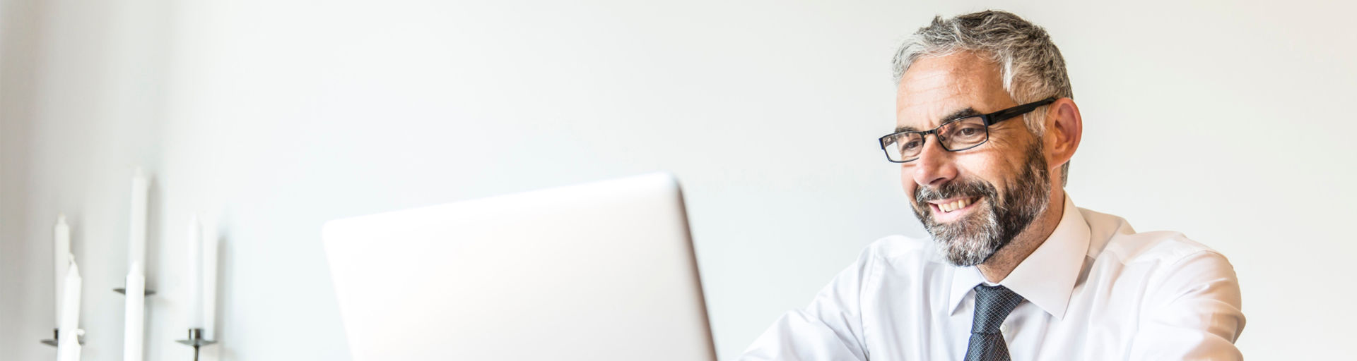 Ein lächelnder Mann mit Brille arbeitet an einem Laptop in einem hellen Raum.