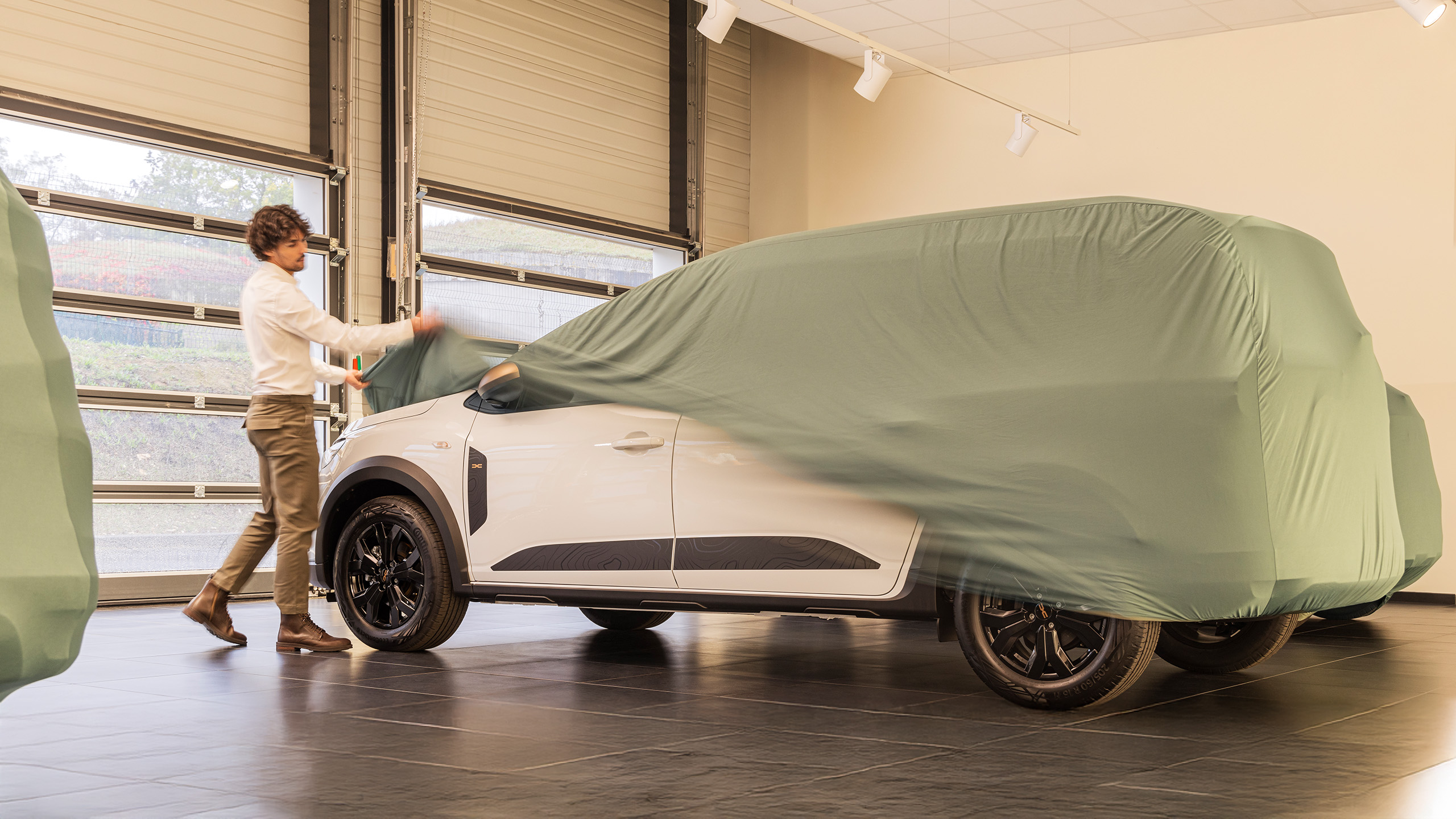 Eine Person zieht eine Abdeckung von einem weißen Auto in einem Autohaus mit großen Fenstern zurück.