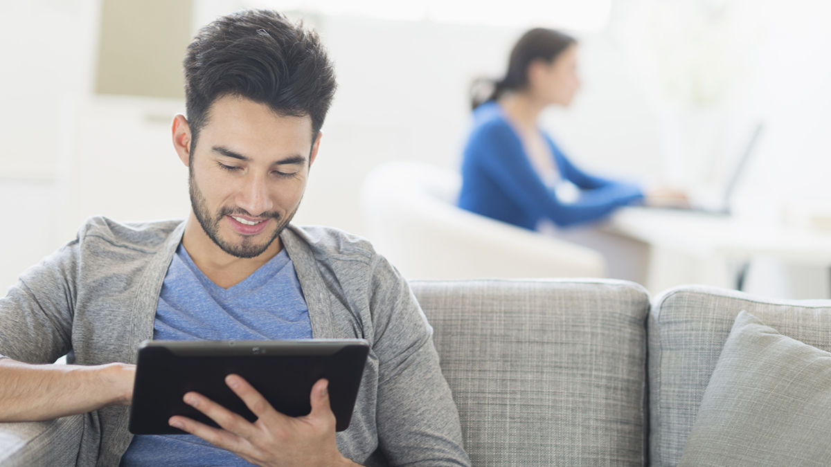 Ein Mann sitzt auf einem Sofa und nutzt ein Tablet, während im Hintergrund eine Frau am Computer arbeitet.