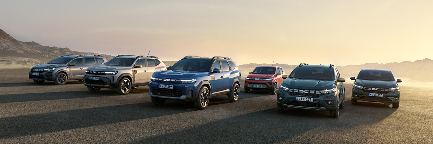 Sechs Dacia SUVs in einer Reihe auf einem sonnigen Weg, mit Bergen im Hintergrund und einer sanften Lichtstimmung.