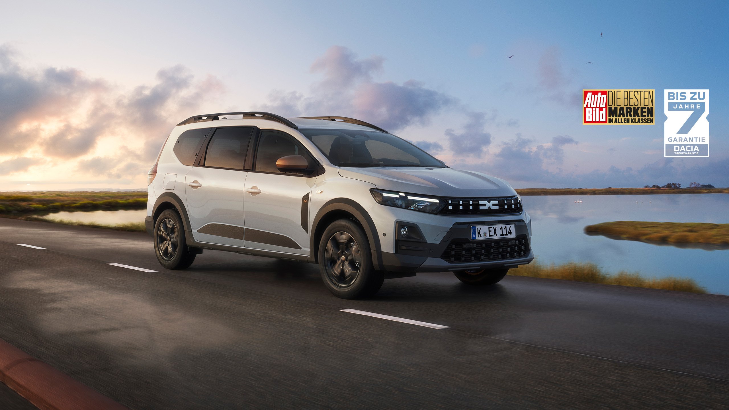 Ein silberner Dacia Duster steht am Strand in der Abenddämmerung. Garantiesymbol unten links.