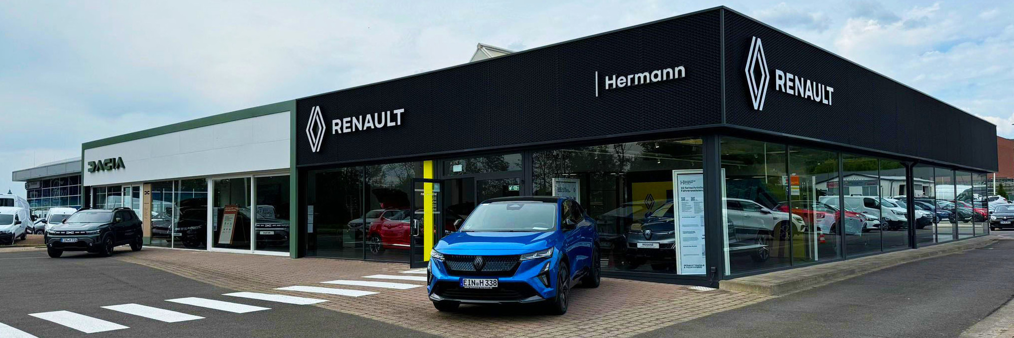 Renault-Händler Hermann mit blauen Auto vor modernem Verkaufsgebäude und ausgestellten Fahrzeugen.