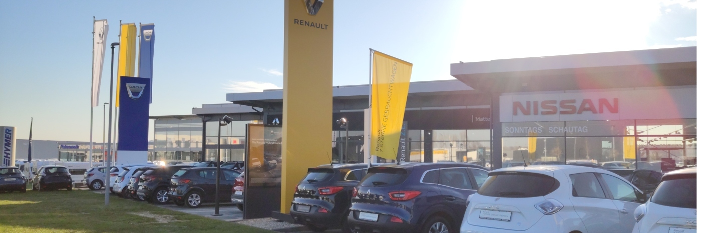 Renault und Nissan Autohaus mit verschiedenen Fahrzeugen und Flaggen im Vordergrund an einem sonnigen Tag.