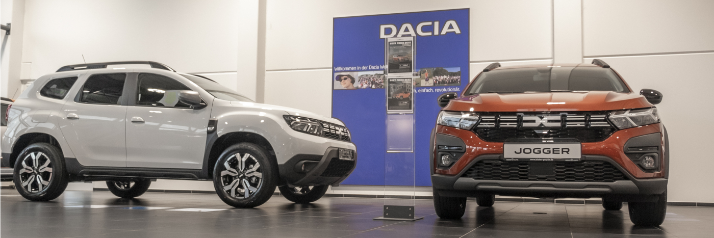 Zwei Dacia Autos, ein weißer SUV und ein orangefarbener Jogger, in einem Ausstellungsraum.