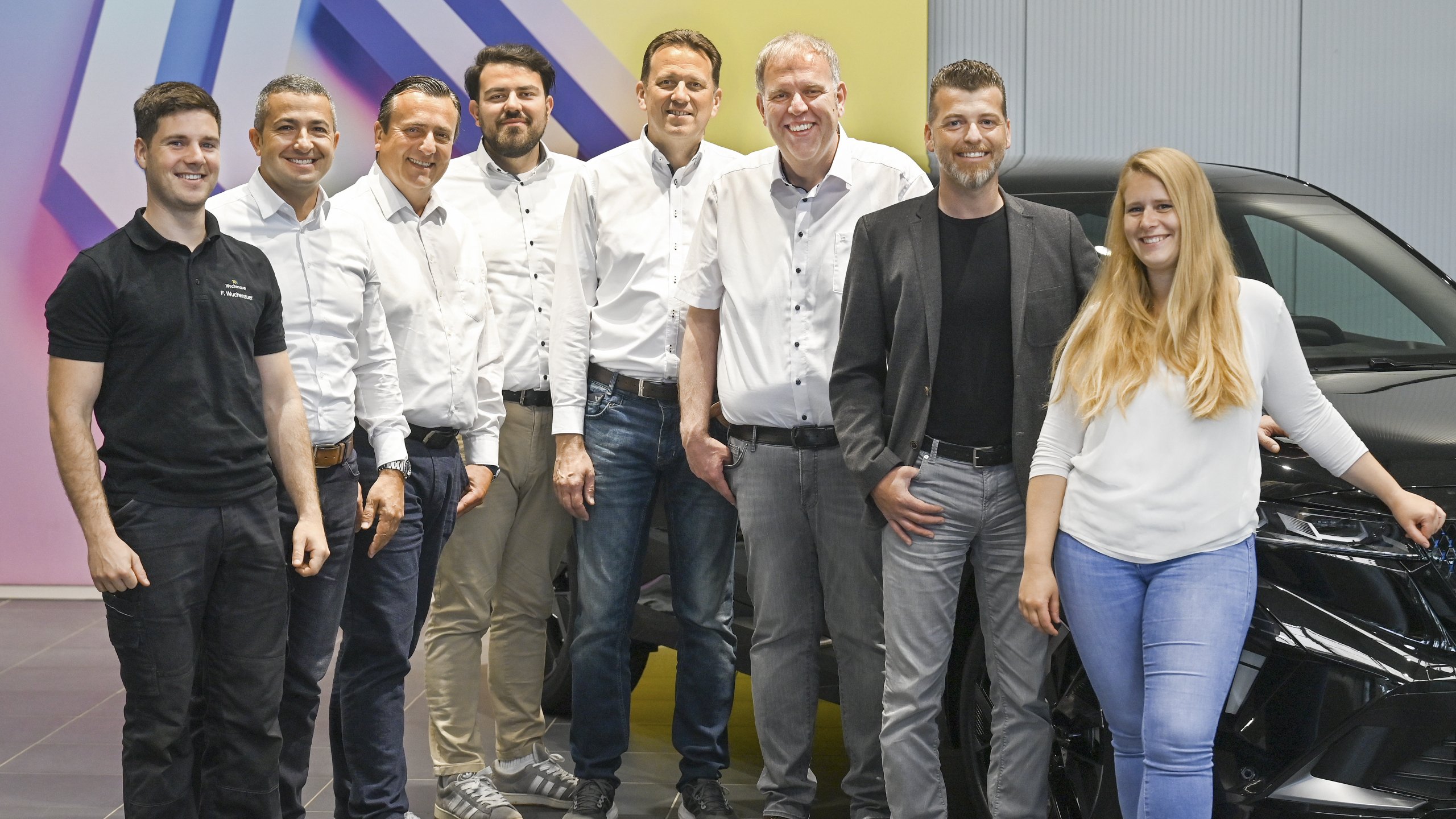 Eine Gruppe von sieben Personen in weißen Hemden und zwei Personen in T-Shirts stehen vor einem Auto und einem bunten Hintergrund.