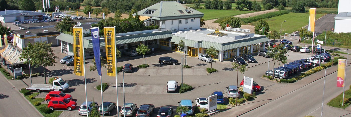 Luftaufnahme eines Autohauses mit Parkplatz, Fahrzeugen und Flaggen in einer grünen Umgebung.