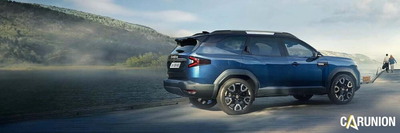 Blauer SUV steht am Ufer eines Gewässers, umgeben von Natur, mit Berglandschaft im Hintergrund.