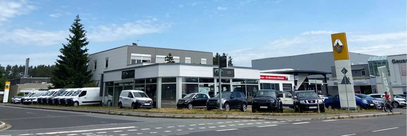 Autohaus mit verschiedenen Fahrzeugen und Renault-Logo, umgeben von Bäumen, an einer Straße.