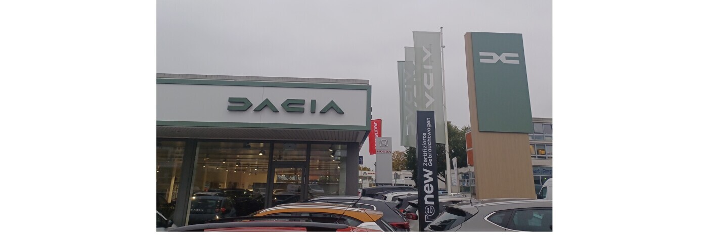 Autohaus mit Dacia-Schild und weiteren Markenlogos, grauer Himmel, geparkte Autos im Vordergrund.