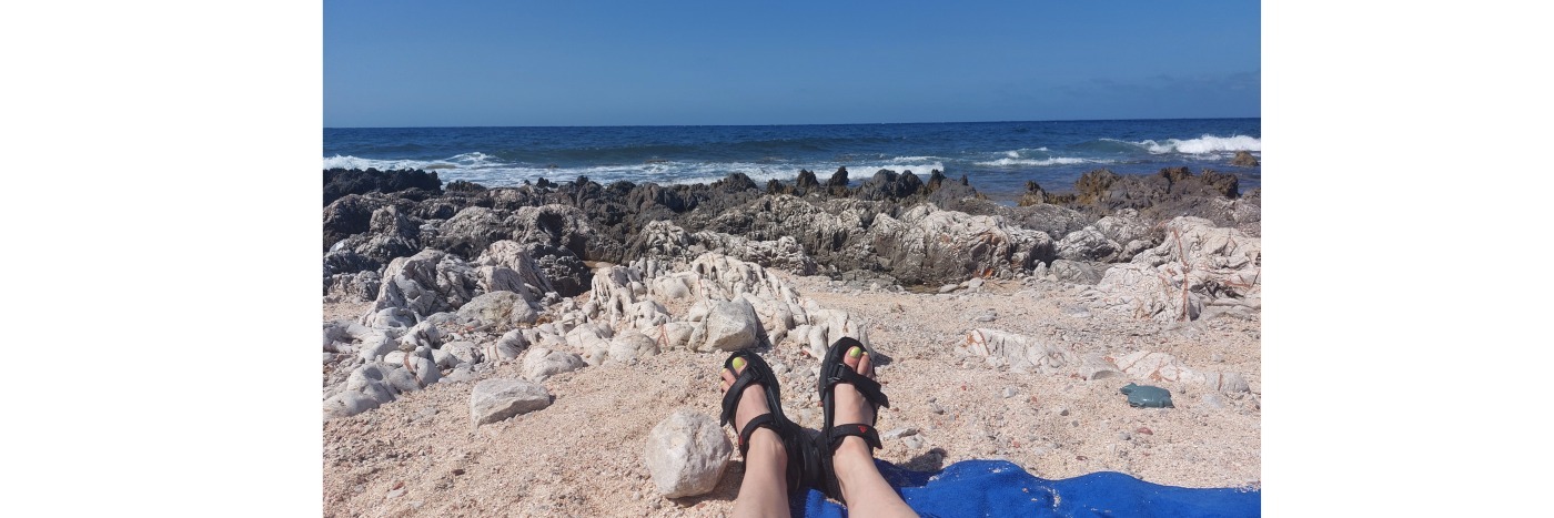 Ein Paar Füße in Sandalen liegen am Strand mit Blick auf das Meer und Felsen im Hintergrund.
